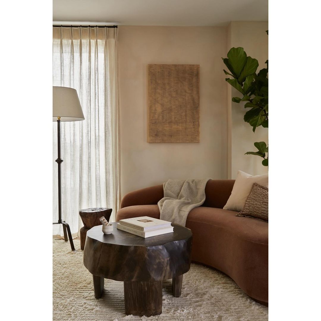 Living room interior with a brown curved sofa, a unique wooden coffee table, a floor lamp, and neutral decor, creating a warm, inviting space.