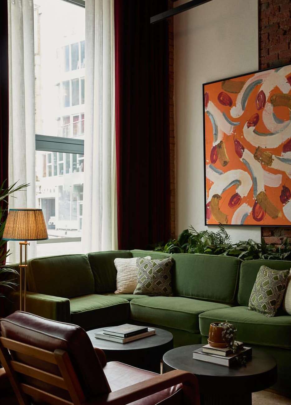 Interior of a hotel lobby with a green velvet corner sofa, patterned pillows, dark wood tables with books, and a colorful painting.