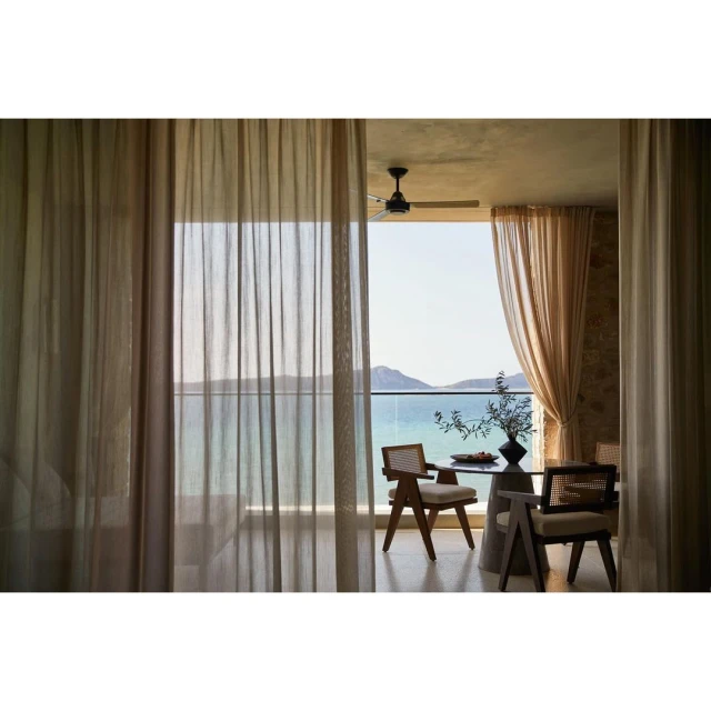 Indoor dining area with woven chairs and a round table on a balcony overlooking a calm ocean view, framed by sheer curtains.