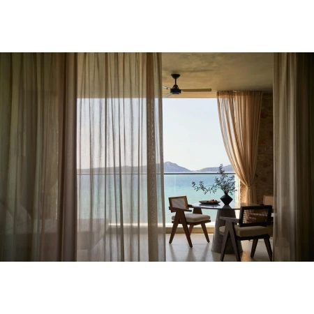 Indoor dining area with woven chairs and a round table on a balcony overlooking a calm ocean view, framed by sheer curtains.