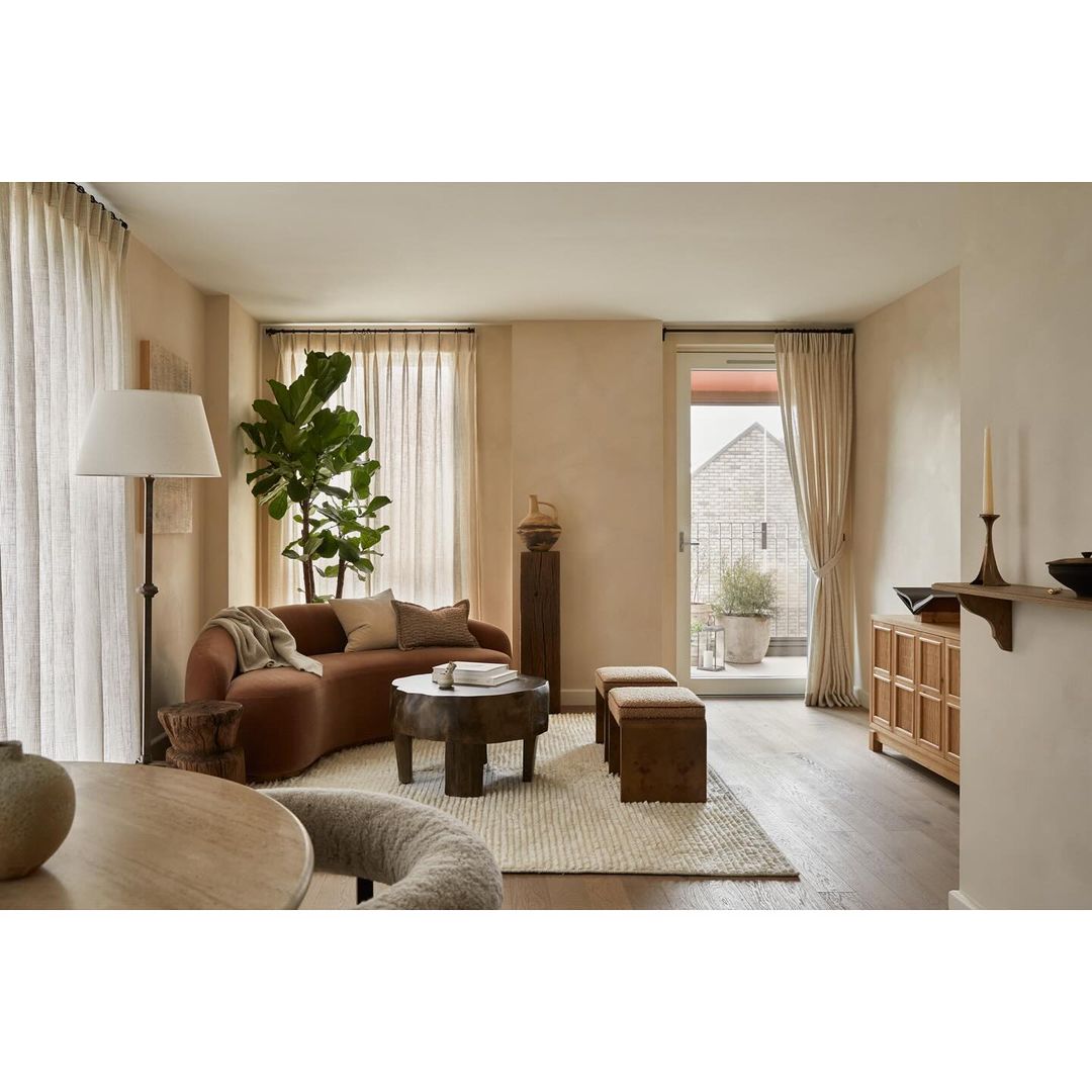 Living room interior with a curved brown sofa, round wood coffee table, benches, potted plant, floor lamp, and neutral color palette.