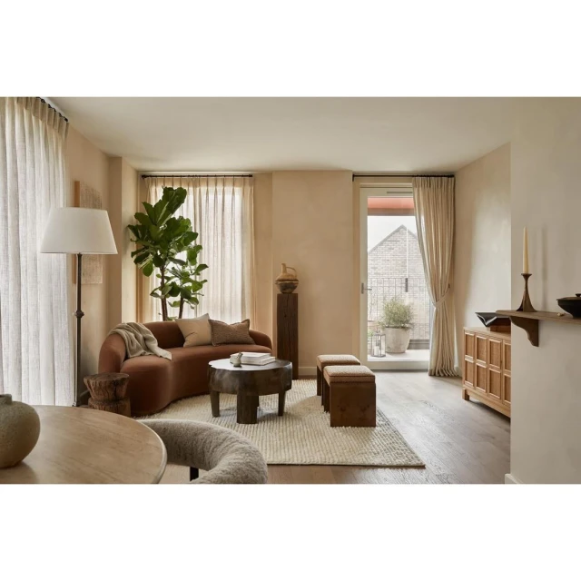Living room interior with a curved brown sofa, round wood coffee table, benches, potted plant, floor lamp, and neutral color palette.