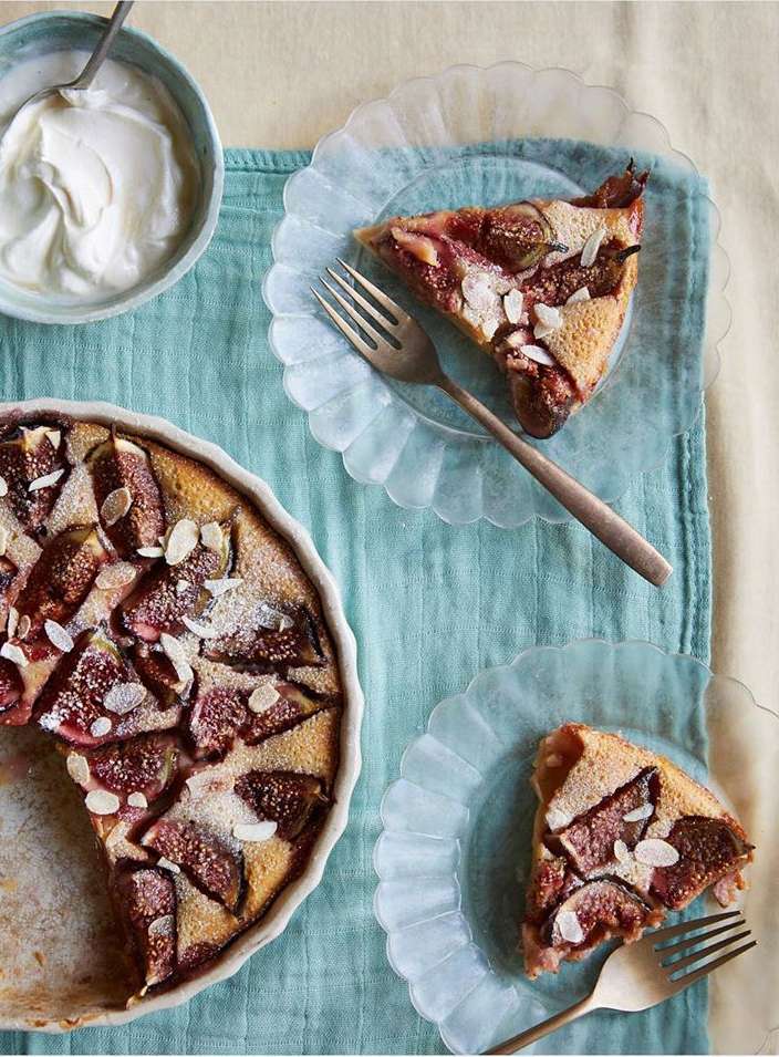 Overhead shot of a fig tart in a white ceramic dish with two slices served on glass plates alongside a bowl of white cream.