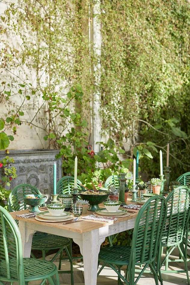 Outdoor dining table is decorated with green and white patterned dishware and candlesticks, surrounded by green wicker chairs.