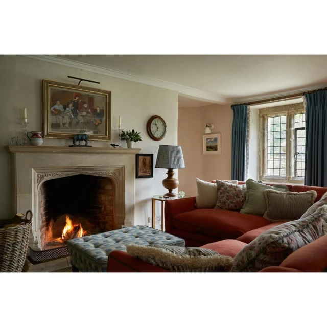 Living room interior with a warm fireplace, comfortable red sofa, patterned ottoman, and soft natural light from a large window.
