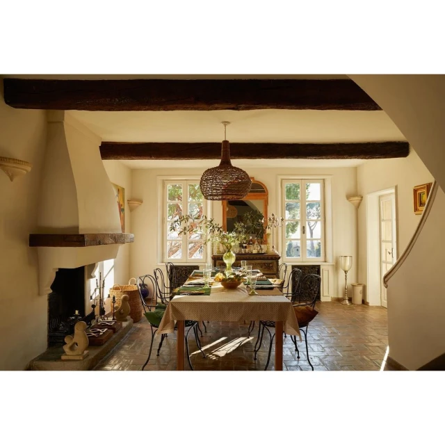 Dining room interior with a table set for a meal, chairs, fireplace, and natural light from windows. Woven light fixture hangs above table.