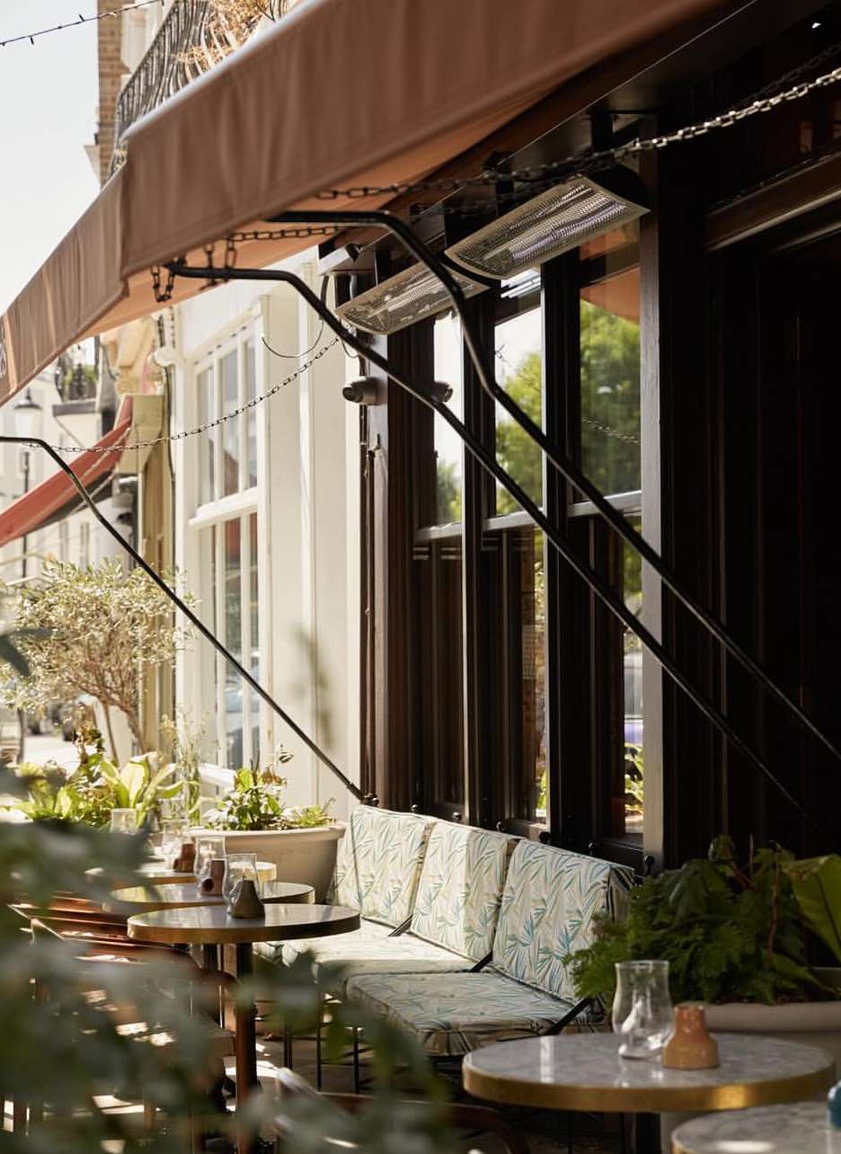 Outdoor seating area with round tables, cushioned benches, and lush greenery, creating an inviting cafe atmosphere in daylight.