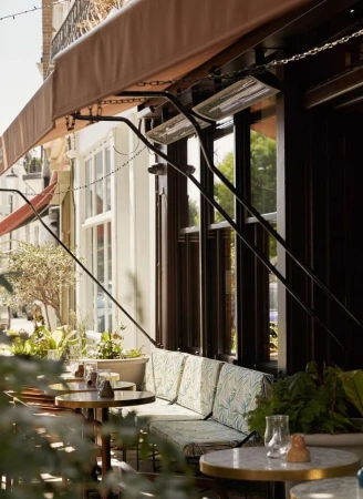 Outdoor seating area with round tables, cushioned benches, and lush greenery, creating an inviting cafe atmosphere in daylight.