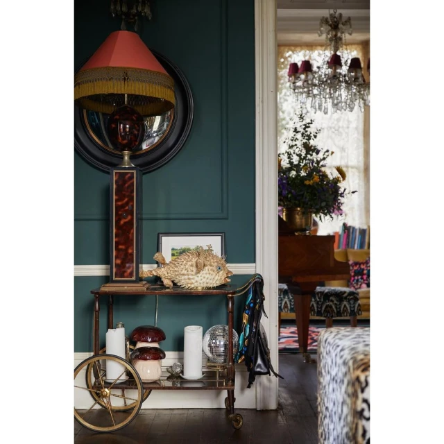 Interior view of a living space featuring a bar cart with decorative items, a floor lamp, and a glimpse into a room with a chandelier.