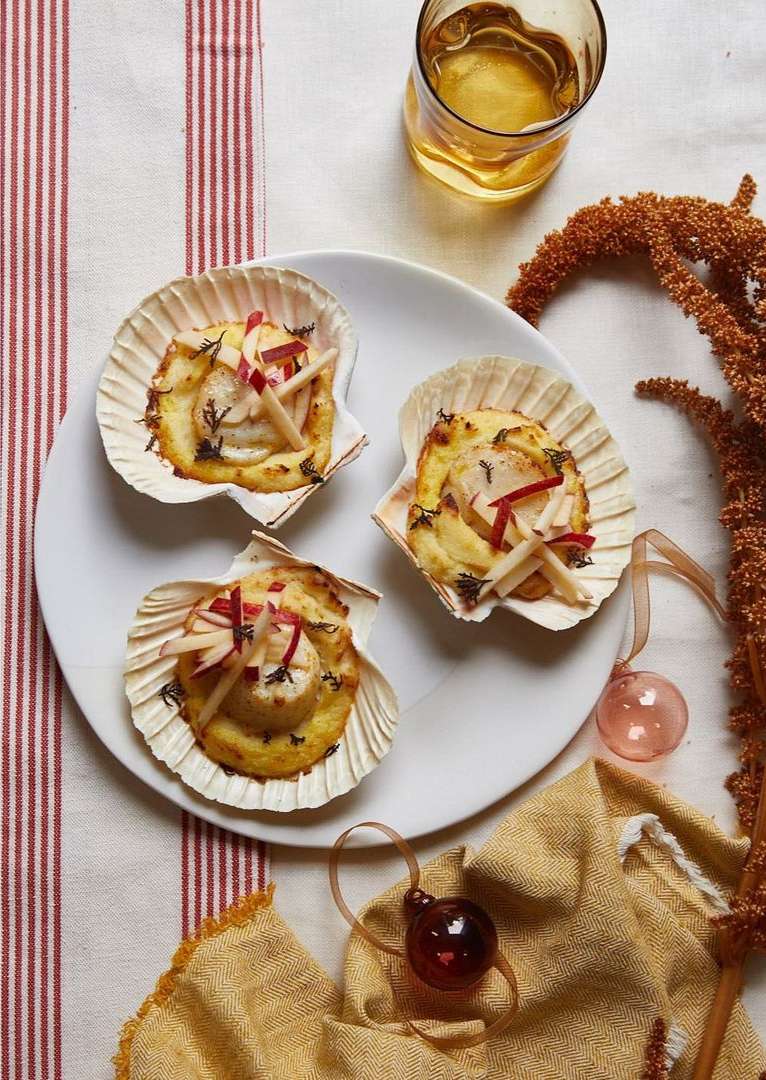 Three scallop shell appetizers on a white plate with a drink, featuring cooked scallops, herbs, and apple matchsticks.