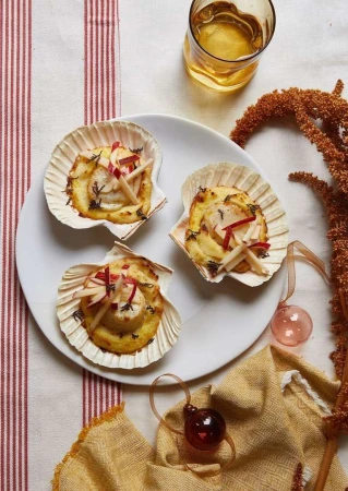 Three scallop shell appetizers on a white plate with a drink, featuring cooked scallops, herbs, and apple matchsticks.