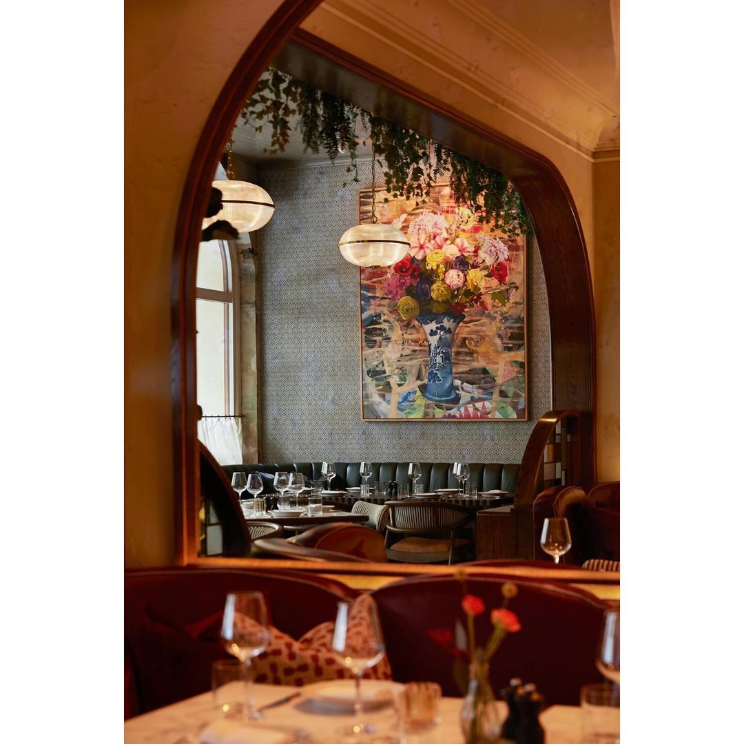 Interior view through a mirror of a restaurant dining room with tables set, floral artwork, and hanging lights in warm lighting.