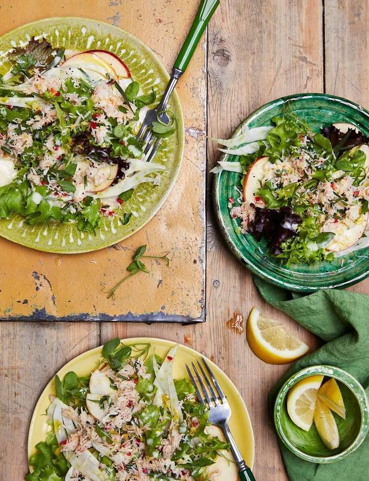 Overhead shot of three plates of salad with tuna, greens, apples, and lemon wedges on a rustic wood table with green accents.
