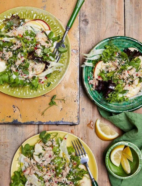 Overhead shot of three plates of salad with tuna, greens, apples, and lemon wedges on a rustic wood table with green accents.