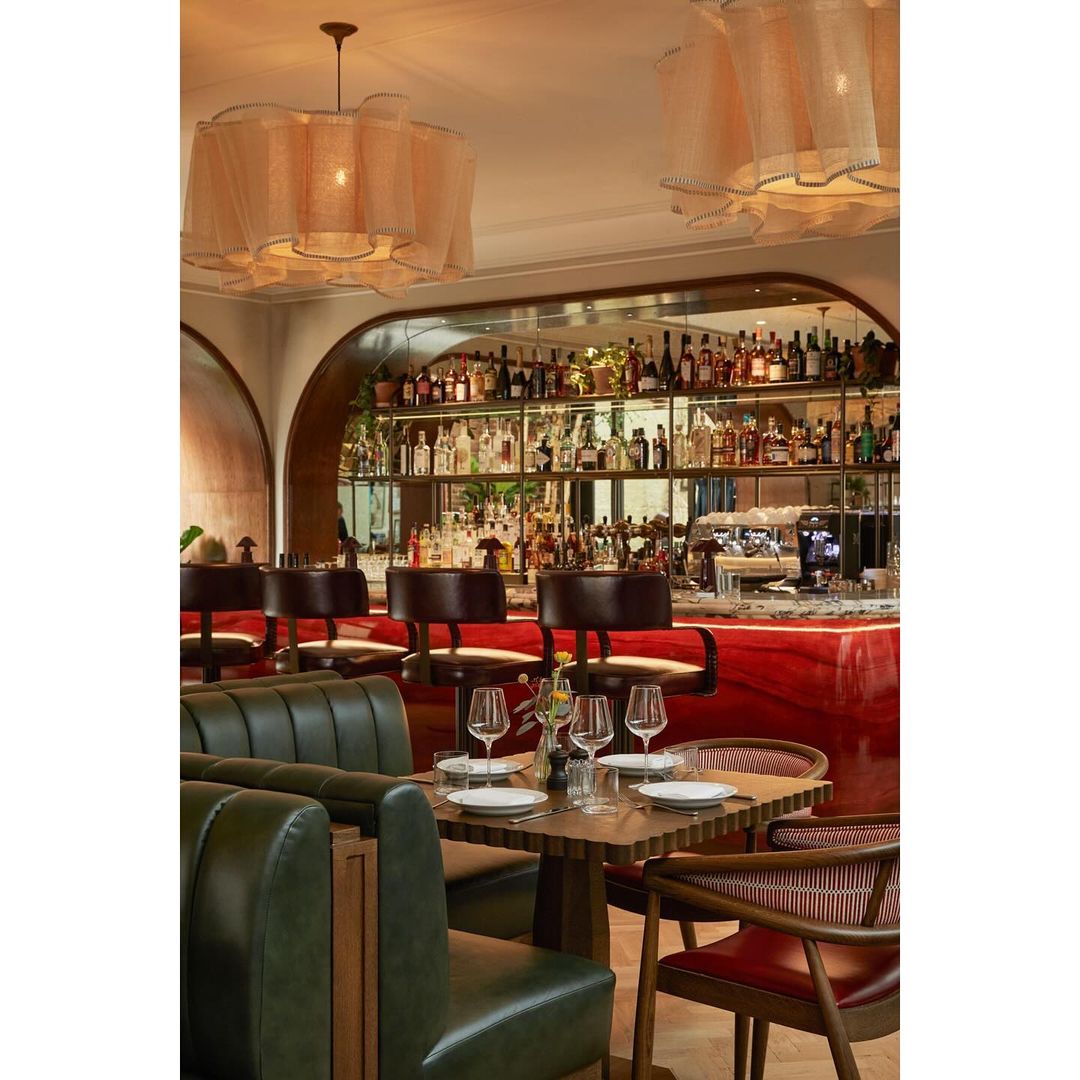 Interior of a restaurant with tables, chairs, booths, overhead lighting, and bar area with bottles in the background.