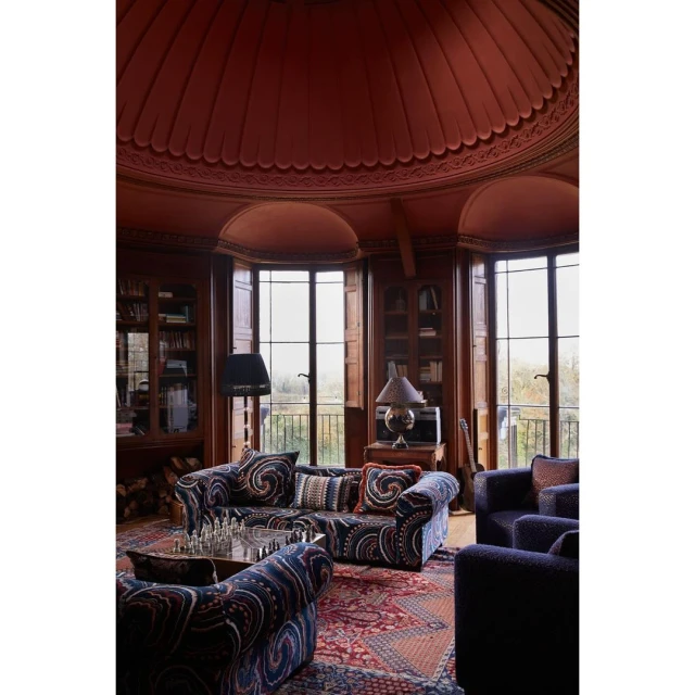 Cozy, ornate living room interior with patterned sofas, bookshelves, dark wood trim, large windows, and a unique domed ceiling.