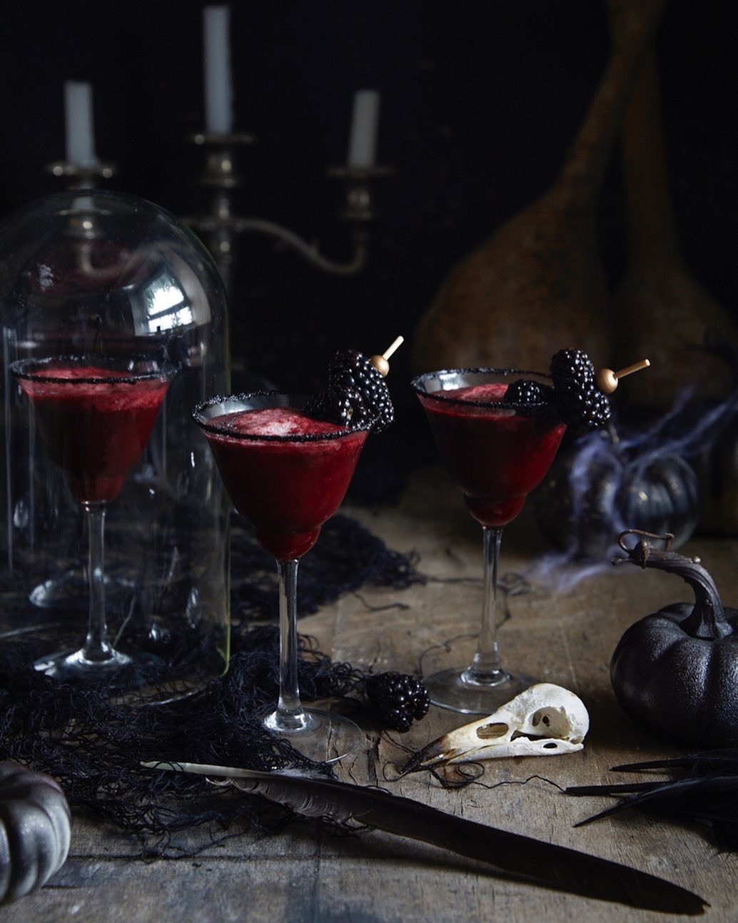 Three red cocktails with blackberries, skull, candles, and spiderwebs on a wooden table against a dark background for Halloween.