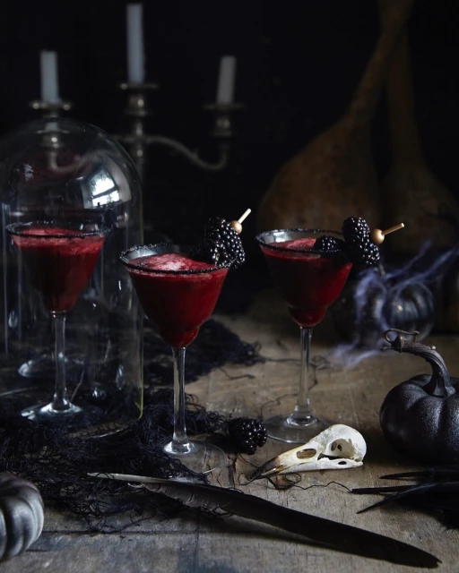 Three red cocktails with blackberries, skull, candles, and spiderwebs on a wooden table against a dark background for Halloween.