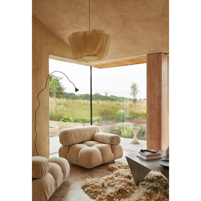 Living room interior with modular sofa, sculptural floor lamp, and view through glass wall to a green field; bright, soft, natural light.
