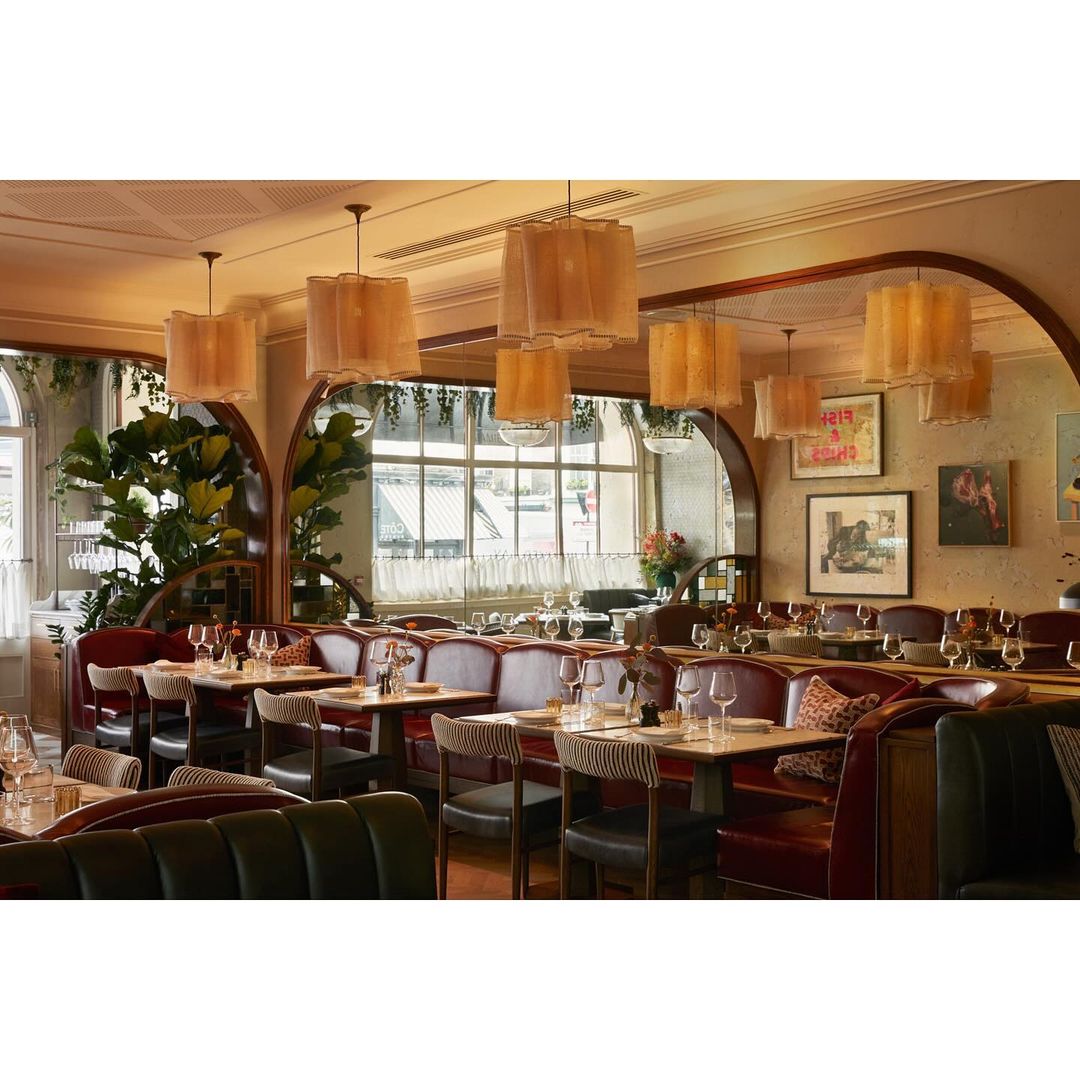 Interior view of a stylish restaurant with booths, tables set with glassware, and modern light fixtures. Plants and artwork decorate the walls.