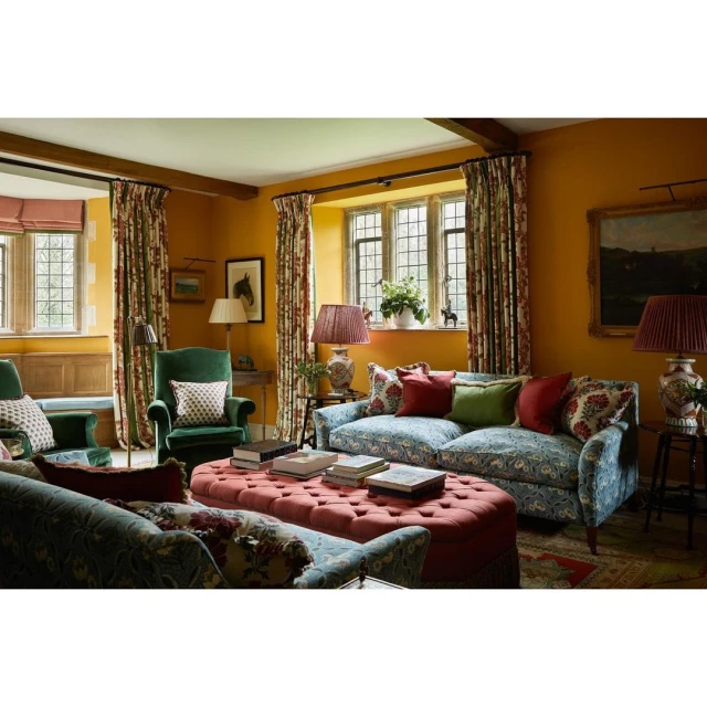 Living room interior with patterned sofas, armchairs, ottoman, floral curtains, artwork, lamps, and natural light from large windows.