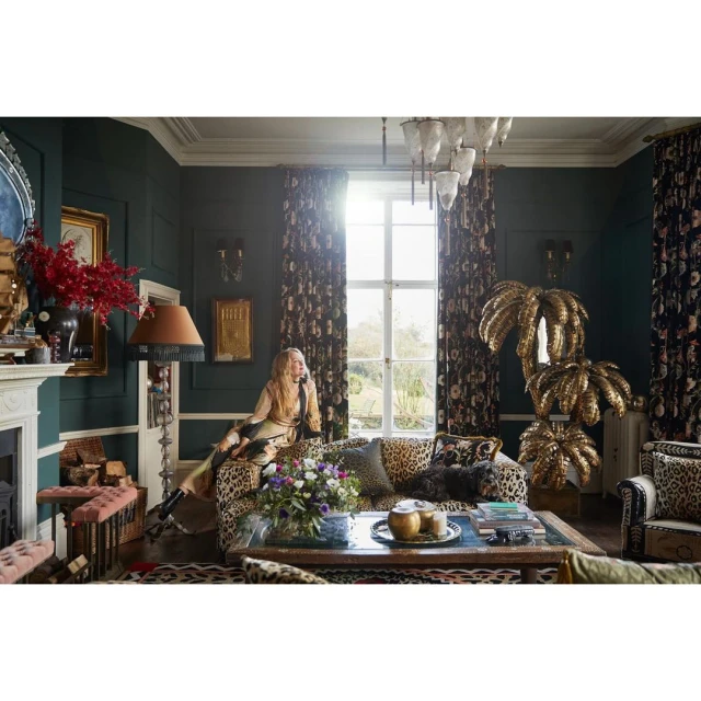 Woman sits on leopard-print couch in luxuriously decorated room featuring floral wallpaper, gold accents, fireplace, and natural light.