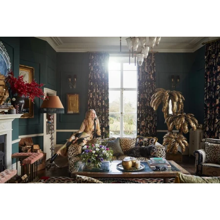Woman sits on leopard-print couch in luxuriously decorated room featuring floral wallpaper, gold accents, fireplace, and natural light.
