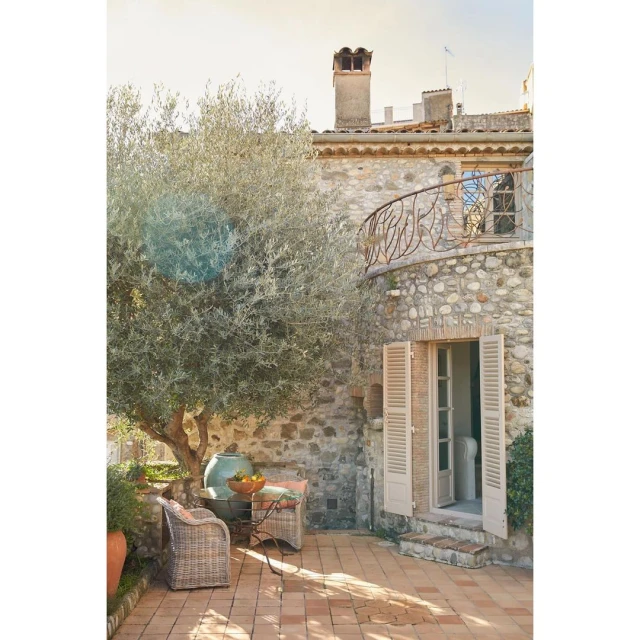 Outdoor patio with a stone building, olive tree, terracotta tiles, and wicker furniture creates a Mediterranean architectural scene.