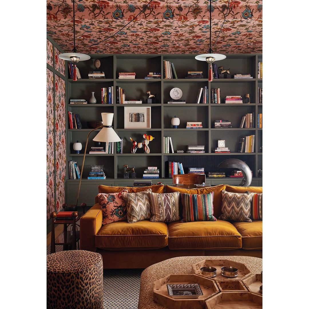 Interior shot of a living room featuring a mustard velvet sofa against built-in green bookshelves and floral wallpaper.