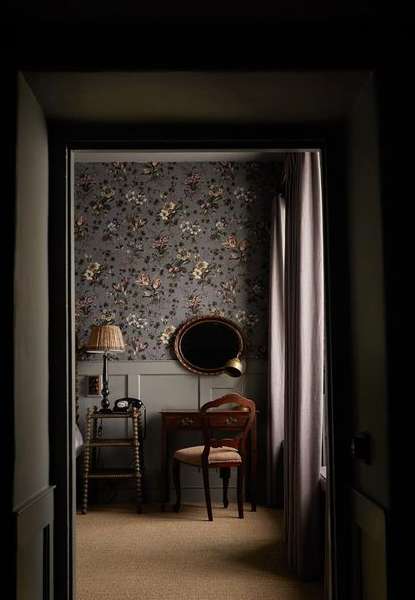 Vintage hotel room interior with floral wallpaper, writing desk and chair, table lamp, and old-fashioned telephone viewed through doorway.