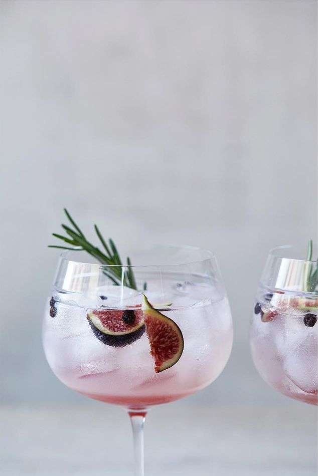 Two clear glasses filled with pink cocktails, ice, figs, and rosemary sprigs, stand against a gray background. Close-up.
