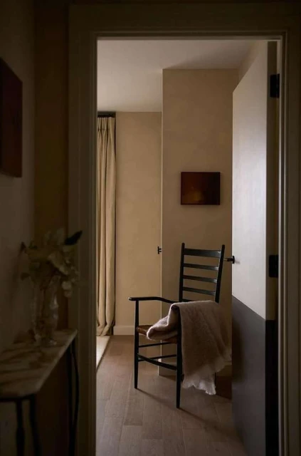 Interior view through doorway: wooden chair draped with a blanket, small painting on the wall, minimal design, in a neutral color palette.