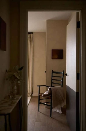 Interior view through doorway: wooden chair draped with a blanket, small painting on the wall, minimal design, in a neutral color palette.