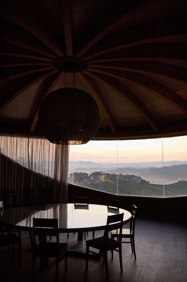 Interior dining room with a round table and chairs looking out a window with a view of rolling hills at dusk.
