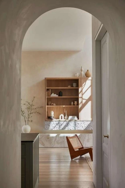 Interior of a living space featuring a light wood bookshelf, chair, and vase with branches, viewed through an arched doorway.