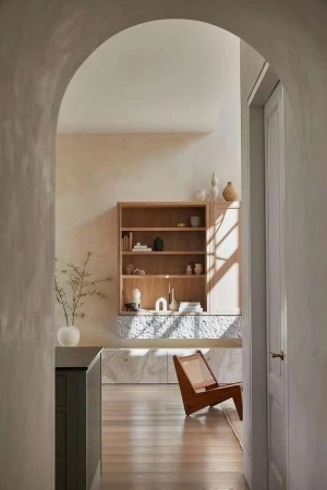 Interior of a living space featuring a light wood bookshelf, chair, and vase with branches, viewed through an arched doorway.