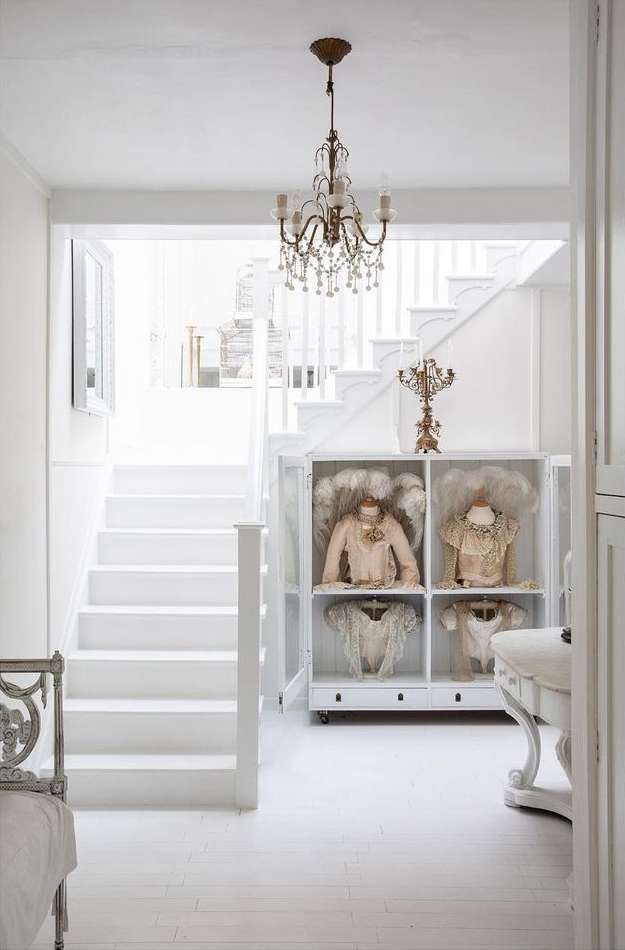 Brightly lit hallway featuring an elegant chandelier, white staircase, and glass cabinet showcasing dresses, creating a clean, chic aesthetic.