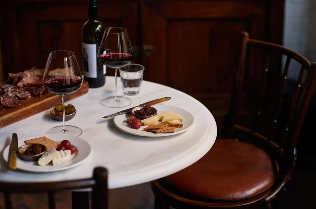 A round marble table is set with wine glasses filled with red wine, cheese, crackers, and charcuterie, presenting a cozy, inviting scene.
