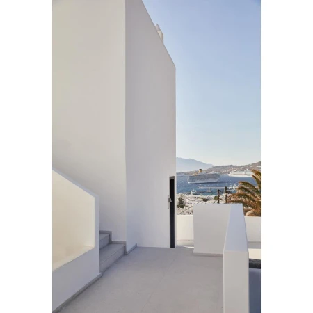 Exterior architecture featuring a white building with stairs and a balcony overlooking the ocean with cruise ships under a clear blue sky.