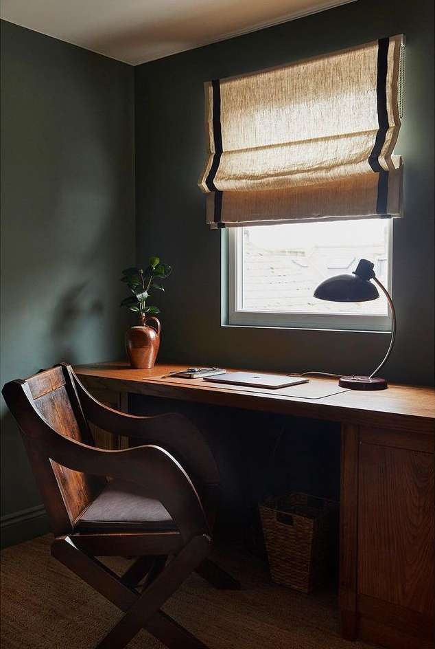 Home office interior with a wooden desk, chair, lamp, and plant, lit by natural light through a window with a tan linen shade.