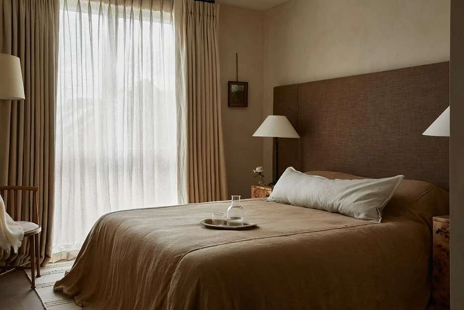 Bedroom interior featuring a made bed with neutral linens, bedside tables with lamps, and soft window light through cream-colored curtains.