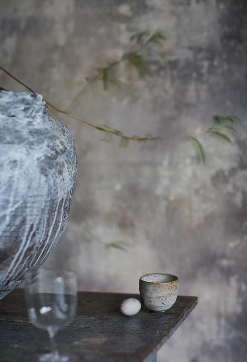 Large textured ceramic vessel with white glaze, small ceramic cup, egg and wine glass on a dark rustic table against a weathered wall.