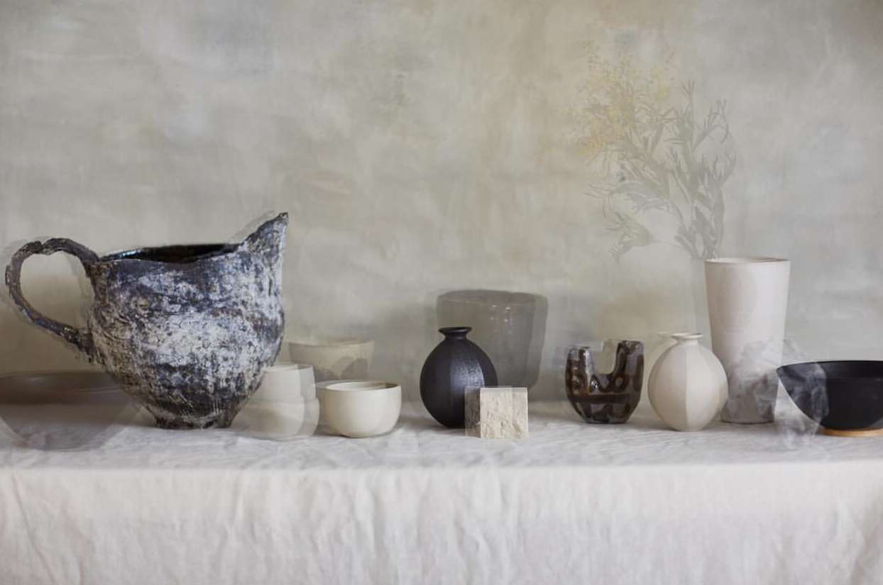 Arrangement of various ceramic vases and bowls in neutral tones and textures on a white tablecloth against a muted background.