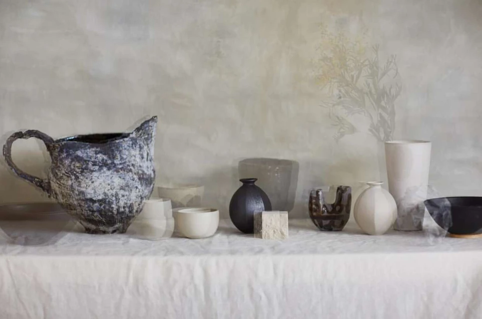 Arrangement of various ceramic vases and bowls in neutral tones and textures on a white tablecloth against a muted background.