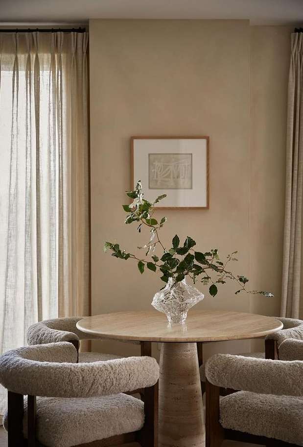 Indoor dining area features round table with a clear vase of greenery, surrounded by four cozy, cream-colored upholstered chairs. Neutral tones dominate.