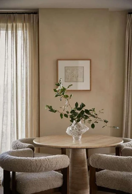 Indoor dining area features round table with a clear vase of greenery, surrounded by four cozy, cream-colored upholstered chairs. Neutral tones dominate.