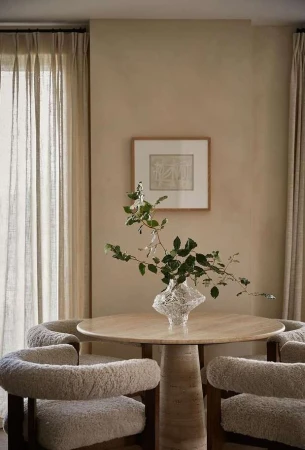 Indoor dining area features round table with a clear vase of greenery, surrounded by four cozy, cream-colored upholstered chairs. Neutral tones dominate.