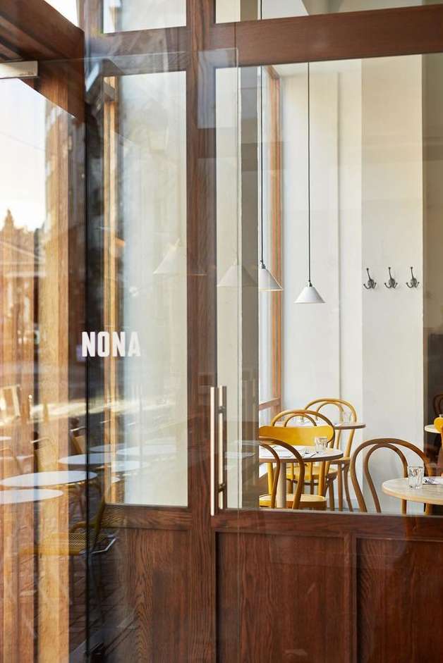 Exterior of Nona restaurant through glass door with wooden frame showing interior with chairs, tables, and hanging pendant lights.