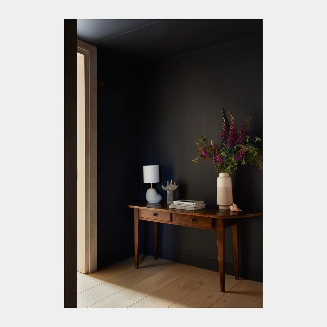 Dark blue interior with wooden table holding flowers, books, and a lamp, featuring a doorway and light wooden flooring.