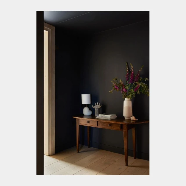 Dark blue interior with wooden table holding flowers, books, and a lamp, featuring a doorway and light wooden flooring.